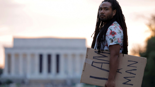 Aaron Xavier Wilson poses for a photo on the National Mall in Washington May 11, 2021, holding the same banner he carried during a "Get Your Knee Off Our Necks" march in support of racial justice last year. Catholic and other faith leaders marked the "painful first anniversary" of George Floyd's May 25, 2020, death with prayers for his family and for "healing, mercy, justice and change." (CNS photo/Andrew Kelly, Reuters)